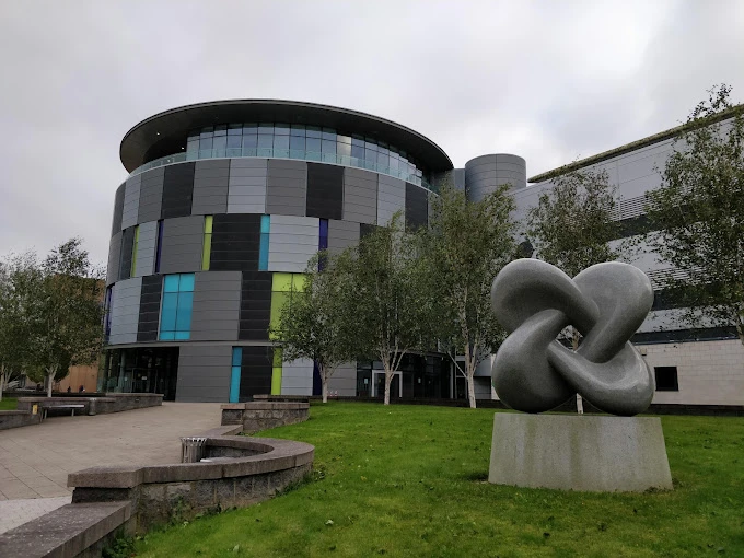 Durham Üniversitesi Modern Kampüs Binası ve Sanatsal Heykel Durham Üniversitesi, modern mimarisi ve sanatsal detaylarıyla dikkat çeken kampüs alanıyla öğrencilere ilham verici bir eğitim ortamı sunuyor. Fotoğrafta, yuvarlak hatlara sahip cam ve panel kaplamalı çağdaş üniversite binası ile ön planda yer alan soyut tasarımlı heykel, çevresindeki yeşil alan ve ağaçlarla birlikte görülmektedir.