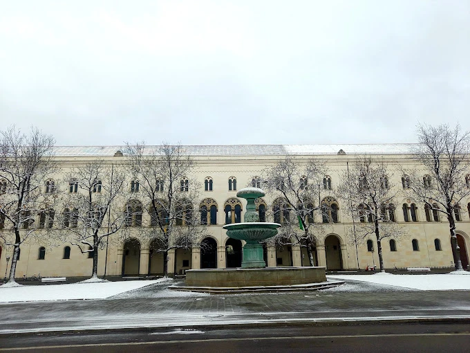 Münih Ludwig Maximilian Üniversitesi Tarihi Binası ve Kış Manzarası Münih Ludwig Maximilian Üniversitesi, Almanya’nın en köklü eğitim kurumlarından biri olarak dikkat çeker. Fotoğrafta üniversitenin tarihi binası önünde yer alan büyük çeşme ve karla kaplanmış kampüs alanı görülmektedir. Kışın beyaz örtüsüyle birleşen bu mimari yapı, hem tarihi hem de estetik açıdan şehrin simgelerinden biridir.