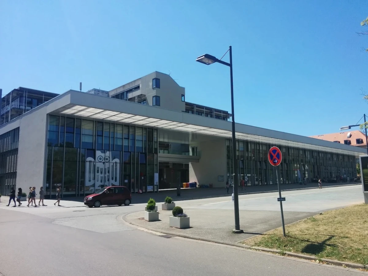 Saarland Üniversitesi Modern Kampüs Binası Saarland Üniversitesi, Almanya’da teknoloji, bilim ve araştırma alanındaki güçlü akademik çalışmalarıyla tanınan prestijli bir kurumdur. Fotoğrafta görülen modern kampüs binası, cam cepheli tasarımı ve ferah giriş alanıyla öğrenciler ve ziyaretçilere çağdaş bir atmosfer sunuyor.