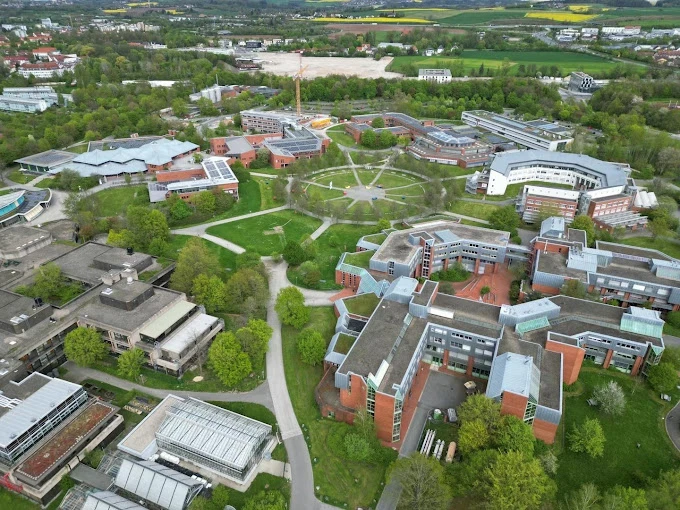 Bayreuth Üniversitesi Yeşil Kampüs Görünümü Bayreuth Üniversitesi, geniş yeşil alanları ve modern mimarisiyle Almanya’nın öne çıkan üniversitelerinden biridir. Fotoğrafta, üniversitenin geniş ve düzenli kampüsü havadan görüntülenmiş; kırmızı tuğlalı binalar, çevresindeki yürüyüş yolları ve yemyeşil alanlarla dikkat çekiyor.