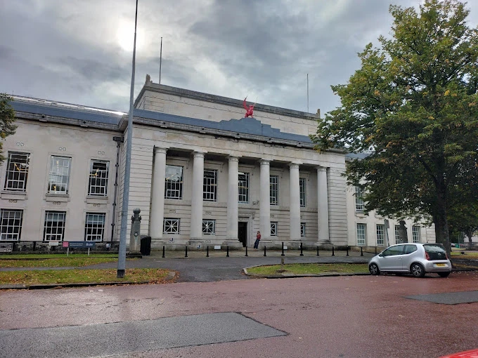 Cardiff Üniversitesi, klasik mimarisi ve görkemli sütunlu giriş cephesiyle dikkat çeken tarihi ana binasıyla öne çıkmaktadır. Fotoğrafta, beyaz taş kaplamalı bina önünde yer alan geniş sütunlar ve çatısında kırmızı ejderha figürü görülmektedir. Bulutlu gökyüzü ve çevredeki ağaçlar, üniversitenin kültürel mirasını ve akademik atmosferini tamamlayan etkileyici bir manzara sunmaktadır.