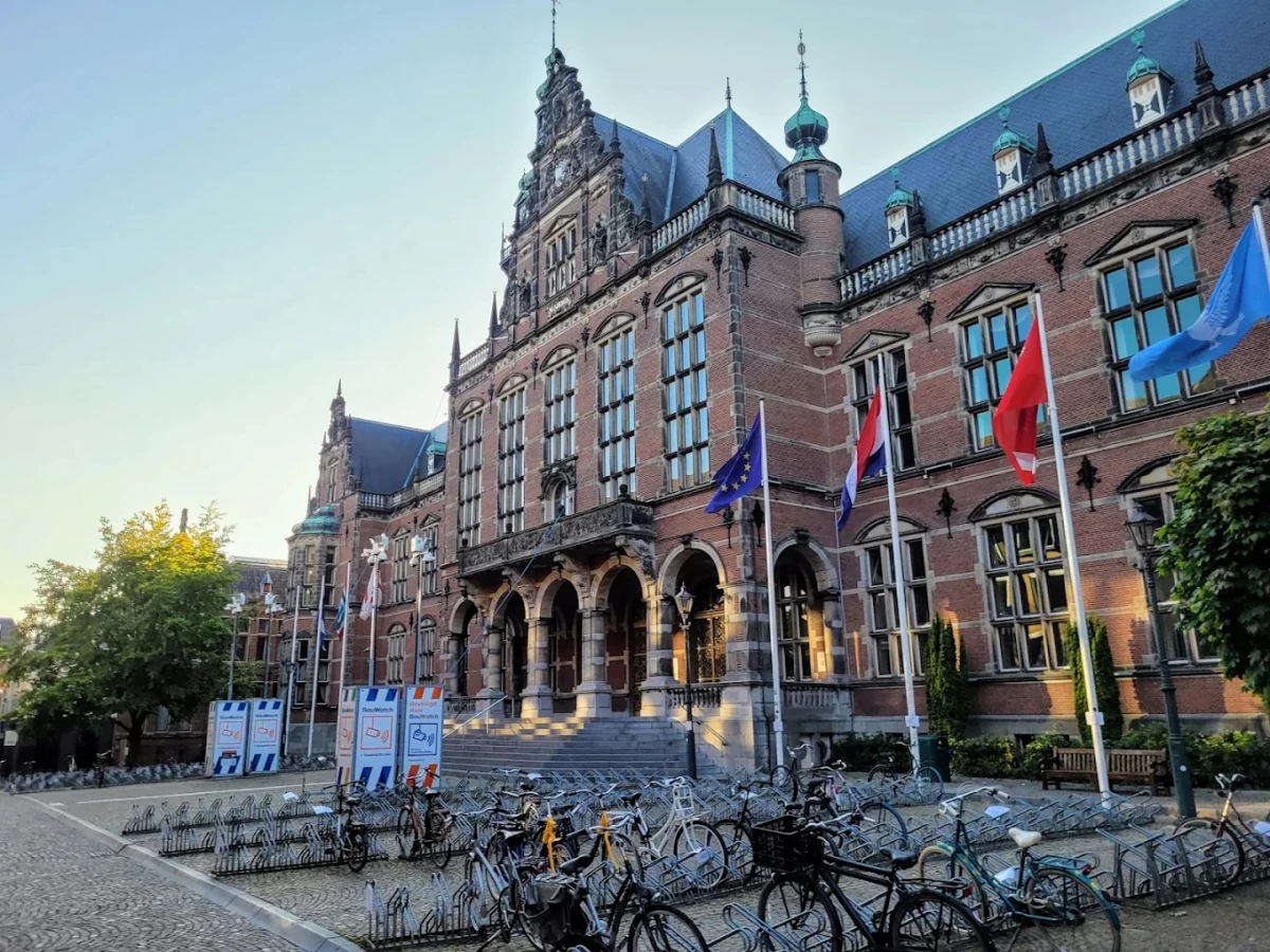 Groningen Üniversitesi Tarihi Ana Bina ve Kampüs Atmosferi Groningen Üniversitesi, Hollanda’nın en köklü eğitim kurumlarından biri olarak tarihi mimarisiyle öne çıkmaktadır. Fotoğrafta, üniversitenin görkemli kırmızı tuğla ana binası, Avrupa Birliği ve ülke bayrakları ile birlikte görülmektedir. Ön planda ise bisiklet park alanı, Groningen Üniversitesi’nin öğrenci dostu ve canlı kampüs yaşamını yansıtmaktadır.